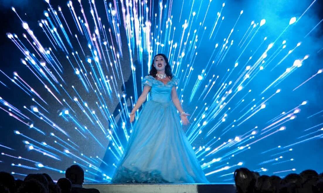 Spektakuläre Szene aus Evita: Eine Sängerin im eleganten blauen Kleid steht im Rampenlicht, während ein funkelndes Lichtspektakel hinter ihr explodiert.
