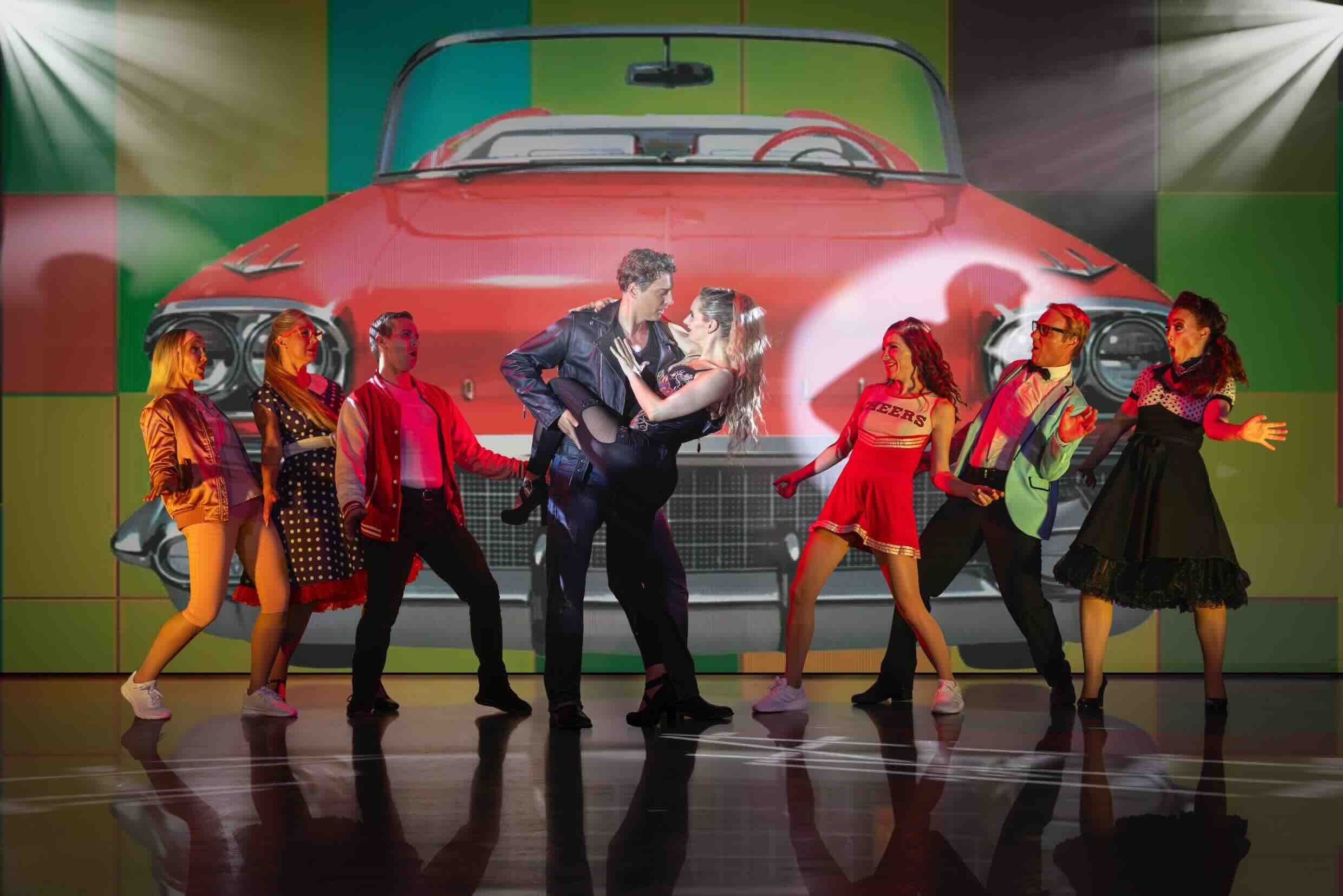 Dynamische Grease-Szene: Ein Paar in Lederjacke und Rock-Outfit im leidenschaftlichen Tanz, umgeben von fröhlichen Tänzern in typischen 50er-Jahre-Looks – vor knallbunter Kulisse mit rotem Oldtimer!